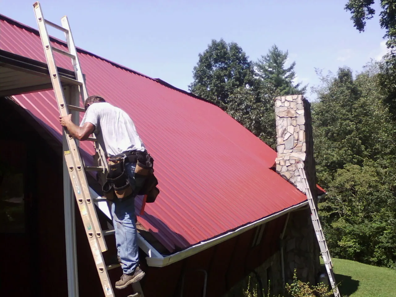 Roofer on a ladder beside a red metal roof and stone chimney — Soffit & Fascia Repair by Roofing Brothers in Atomic City